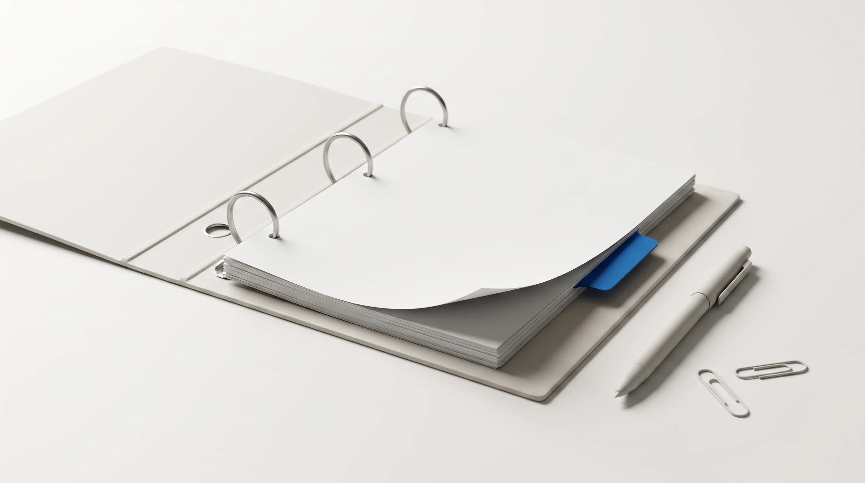 Open ringband met blanco vellen en één blauw tabblad, met een pen en twee paperclips ernaast op een lichte ondergrond