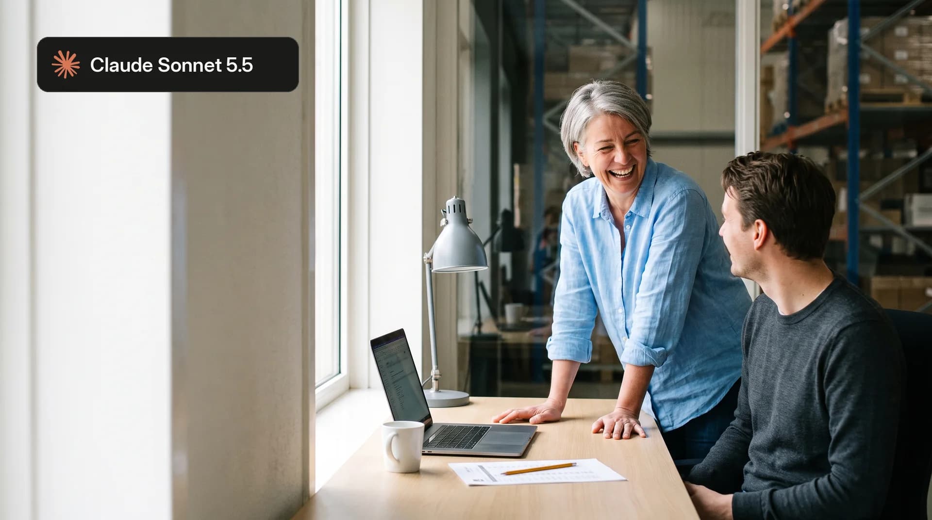 Lachende vrouw bij een bureau met een collega achter een laptop in een magazijnkantoor, met het label Claude Sonnet 5.5