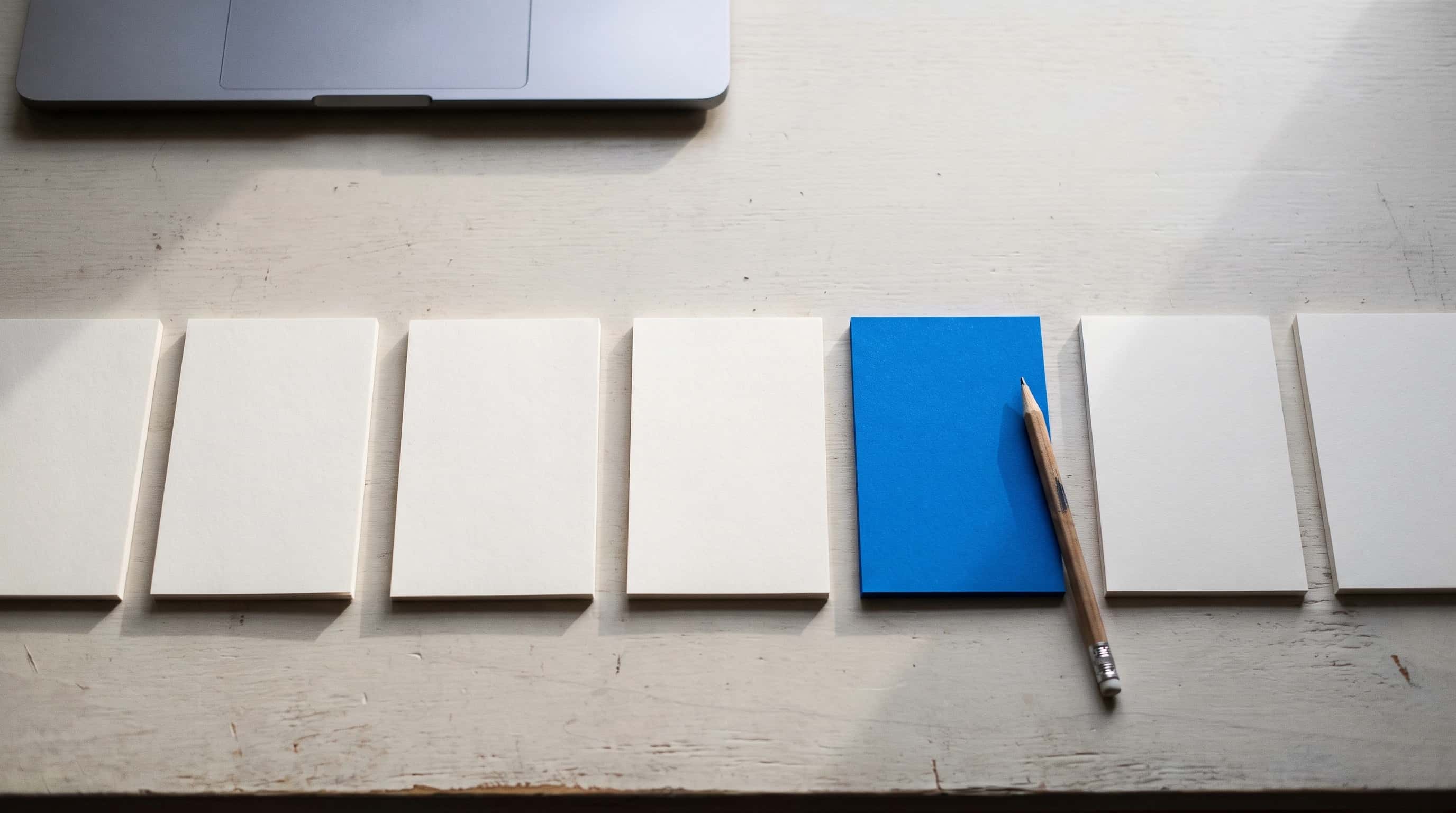 Zeven blanco kaartjes in een rij op een licht bureau, één ervan blauw, met een potlood en de rand van een laptop ernaast