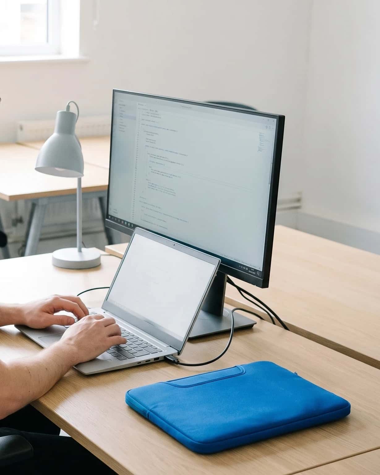Twee handen op een laptop naast een groot scherm met code; op het bureau ligt een blauwe laptopsleeve, daglicht van links.